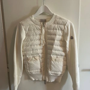 Vit quiltad kofta från Moncler - Snygg vit kofta från Moncler med quiltad framsida och två fickor med knapp. Långärmad, rund hals och dragkedja framtill. Moncler-logga på ärmen. Materialmix med stickade ärmar och rygg samt polyester på framsidan.