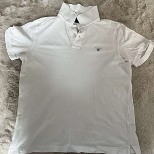 Vit piké t-shirt från Gant, M - Vit piké t-shirt från Gant med klassisk krage och knappar framtill. Broderad Gant-logga på bröstet. Tillverkad i mjuk bomull och har korta ärmar. Perfekt för en clean och stilren look.