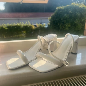 Vita sandaler med klack från XIT - Stilrena vita sandaler från XIT med fyrkantig tå och breda remmar. Skorna har en chunky låg klack och justerbart spänne vid ankeln. Tillverkade i syntetmaterial med platt sula och öppen tå, perfekta för sommarens alla tillfällen.