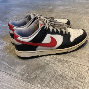 Nike Dunk Low svart/vit/röd - Nike Dunk Low sneakers i svart, vitt och rött med klassisk röd swoosh på sidorna. Ovandel i skinn med perforerad tåbox och grå snörning. Låg modell med bekväm vadderad insida och slitstark yttersula. Perfekta för dig som gillar streetstyle och retro vibes.