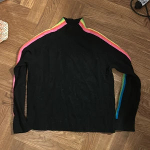 Svart polotröja med regnbågsränder From Future - Svart polotröja från From Future med färgglada regnbågsränder längs axlar och ärmar. Tröjan är stickad och har lång ärm samt ribbade muddar. Perfekt för dig som vill sticka ut med en cool och färgstark detalj. Cashmir