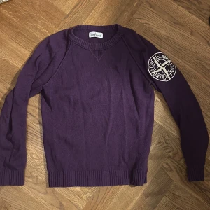 Lila stickad tröja från Stone Island - Snygg lila stickad tröja från Stone Island Junior med rund hals och ribbade muddar. Tröjan har lång ärm och en stor broderad Stone Island-logga på ena ärmen. Bomull