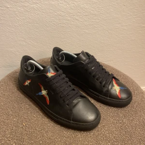 Svarta Axel Arigato sneakers med broderade fåglar - Svarta sneakers från Axel Arigato i skinn med broderade färgglada fåglar på sidorna och framtill. Klassisk låg modell med svarta snören och rund tå. Snygg detalj med broderiet som ger en unik vibe till looken.