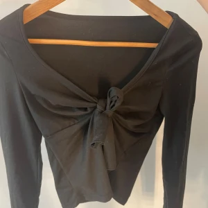 Svart långärmad topp med knyt från SHEIN - Svart långärmad topp från SHEIN i storlek XS. Toppen har en djup v-ringning och snygg knytdetalj framtill som ger en cool och trendig look. Tillverkad i mjukt och stretchigt material som sitter skönt på kroppen. 