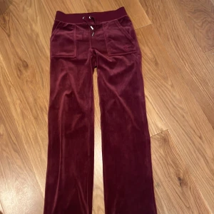 Vinröda velourbyxor Juicy Couture XS - Mysiga vinröda mjukisbyxor från Juicy Couture i velour. Byxorna har bred resår och snörning i midjan, stora fickor fram och bak med knappdetaljer. Perfekta för chill eller en avslappnad look. Jag säljer de för att de bara ligger i min garderob och används inte 