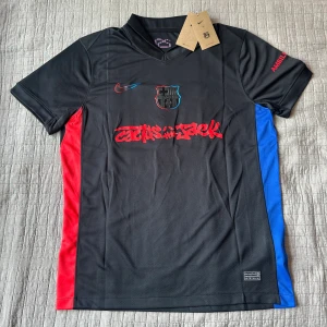 FC Barcelona x Nike Lamine Yamal tröja - Säljer en svart FC Barcelona x Travis scott fotbollströja med röda och blå sidopaneler, Dri-FIT-material och tryck 'Cactus Jack' framtill. Lamine Yamal och nummer 19 på ryggen. Broderat klubbmärke och Nike-logga på bröstet.  Skicka om ni har några funderingar!😁