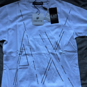 Vit t-shirt från Armani Exchange - Snygg vit t-shirt från Armani Exchange med stora svarta bokstäver och logga tryckt över hela framsidan. Klassisk rund hals och korta ärmar. Perfekt för dig som gillar stilrena och trendiga plagg med tydlig branding.