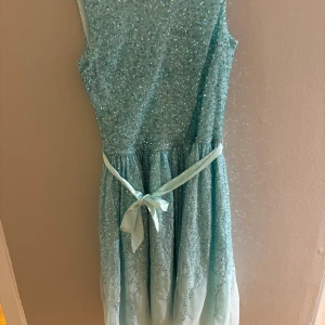 Ljusblå glittrig klänning med rosett - Säljer en ljusblå glittrig klänning med paljetter och ett fint satinband som knyts i midjan. Klänningen är ärmlös och har en lätt, fluffig kjol som ger en drömmig känsla. Perfekt för dig som vill sticka ut med lite extra glitter och glam.