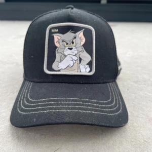Svart Tom & Jerry keps mesh - Svart keps med mesh på sidorna och bak, samt broderad patch framtill med Tom från Tom & Jerry. Grå och vita kontrastsömmar på skärmen och en Tom & Jerry-logga på sidan. Perfekt för dig som gillar retro och tecknade serier.