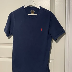 Mörkblå t-shirt från Polo Ralph Lauren - Snygg mörkblå t-shirt från Polo Ralph Lauren med klassisk röd broderad logga på bröstet. Modellen är kortärmad och har en rund hals. Tillverkad i mjuk bomull som känns skön mot huden. Perfekt för dig som gillar stilrena och tidlösa plagg.