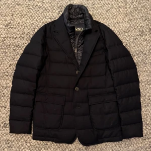 Herno jacka - Svart dunrock från Herno med quiltad design och klassisk kavajkrage. Jackan har två stora fickor framtill, knäppning med två knappar och en innerdel med dragkedja. Materialet är dun och polyester, perfekt för kalla dagar. Snygg och stilren look med modern touch.