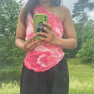 Rosa one shoulder topp i mesh-look - Säljer en snygg rosa och vit one shoulder topp i mesh-liknande polyester. Toppen har ett unikt batikmönster och är ärmlös med asymmetrisk skärning. Perfekt för dig som vill sticka ut med färg och cool form. Tunn och luftig, passar till sommarens alla outfits.