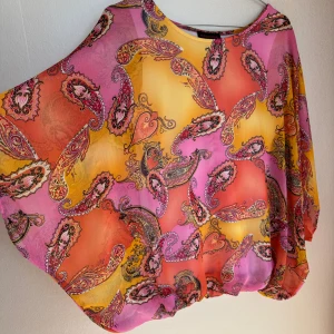 Färgglad blus från Mascara - Säljer en luftig blus från Mascara med paisleymönster i rosa, orange och gult. Blusen har lös passform, rund halsringning och långa, vida ärmar. Materialet är tunt och flowy, perfekt för fest när du vill sticka ut med färg.