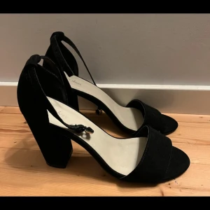 Svarta klackskor  - Stilrena svarta sandaler från Nelly med bred klack och öppen tå. Skorna har en enkel rem över tårna och justerbar ankelrem med spänne. Ytan är i mockaliknande material som ger en lyxig känsla. Perfekta för dig som vill ha en klassisk och trendig look. Använda 1 gång.