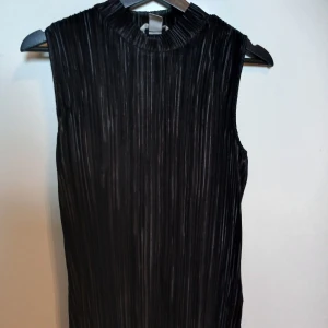 Svart plisserad ärmlös topp - Snygg svart plisserad topp med hög krage och ärmlös design. Materialet har en glansig finish som ger en cool och modern vibe. Perfekt för dig som gillar minimalistisk stil med en twist. Passar dig som vill sticka ut utan att vara för mycket.