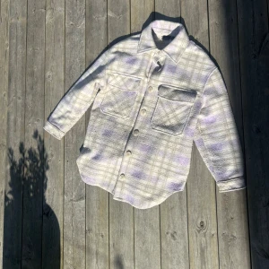 Rutig overshirt från Vero Moda - Säljer en oversized rutig overshirt från Vero Moda i ljusa toner av beige, vit och lila. Skjortan har stora bröstfickor, knappar framtill och lång ärm. Perfekt att slänga över en hoodie eller t-shirt för en chill look.