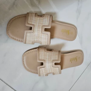 Beige sandaler från Shein - Stilrena beige sandaler från Shein med bred flätad rem i syntetmaterial och öppen tå. Snyggt mönstrad ovandel i ljusa toner och platt sula för en avslappnad look. Perfekta för varma dagar.