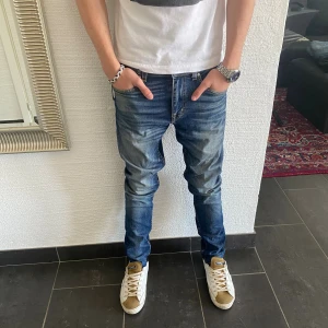 Tiger Of Sweden - Säljer ett par snygga blå jeans från Tiger of Sweden, modell Evolve. Jeansen har klassisk femficksdesign, normal passform och raka ben. Tillverkade i Italien av slitstarkt denim med coola slitningar och tvättade detaljer. Perfekta för en avslappnad och trendig look.