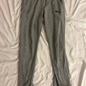 Grå mjukisbyxor från Puma - Snygga grå mjukisbyxor från Puma med svart logga på ena benet. Byxorna har en avslappnad passform och är gjorda i mjukt bomullsmaterial. Perfekta för chill eller träning. Elastisk midja och fickor på sidorna.