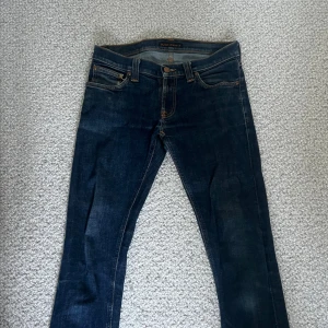 Mörkblå jeans från Nudie  - Extremt feta mörkblåa jeans från Nudie med klassiska orange sömmar och broderade bakfickor. Modellen har normal passform och raka ben. En liten slitning vid vänster ficka som gör dom snyggare och jeansen är i väldigt bra skick. Skriv för minsta lilla fråga eller fundering!