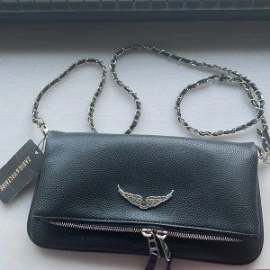 Svart axelväska Zadig & Voltaire skinn - Cool svart axelväska från Zadig & Voltaire i äkta skinn med silverfärgad kedjerem och snyggt vinge-emblem framtill. Väskan har dragkedja både fram och bak. Har tre fack att lägga grejer i. Köpte för 3600 kr. Axelbandet på ena sidan snöra har gått upp lite men syns knappt. 
