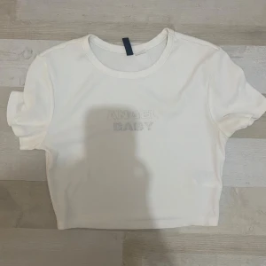 Vit croppad topp med text Angel Baby - Superfin vit croppad topp med korta ärmar och broderad texten 'ANGEL BABY' framtill i glittrigt silver. Ribbad struktur i tyget och rund halsringning. Perfekt för dig som gillar en söt och trendig stil.