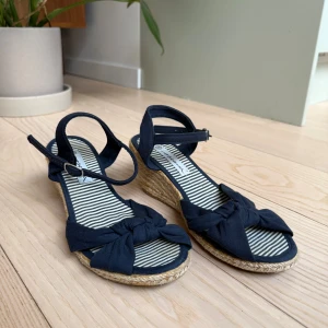 Marinblå kilklack sandaler med rosett - Snygga marinblå sandaler med kilklack i flätad jute och öppen tå. Ovandelen har en knytdetalj framtill och justerbart spänne runt vristen. Har varit mina skönaste klackar att gå i och skaver ingenting då det bara är ett ”tygstycke” runt fötterna. Strlk 38 men passat mig bra också som brukar vara 37 1/3