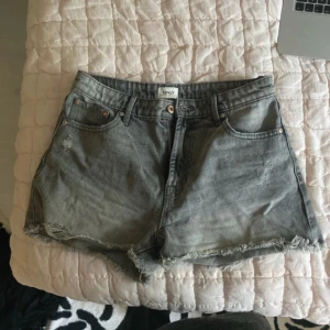 Grå jeansshorts från ONLY - Snygga grå jeansshorts från ONLY med fransig kant och klassisk femficksdesign. Modellen är högmidjad och har dragkedja och knapp framtill. Materialet är slitstarkt denim och shortsen har en cool, lite tvättad look som passar perfekt till sommaren.