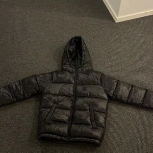 Svart pufferjacka från Grunt, L - Säljer en svart pufferjacka från Grunt i storlek L. Jackan har huva, dragkedja framtill och breda, vadderade paneler som ger en riktigt fet look. Perfekt för kalla dagar när du vill hålla stilen och värmen. Snygg och enkel att matcha med allt. Och perfekt nu till vintern. Prisen går o snacka om vid snabb affär.