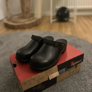 Svarta träskor från Sievi, stl 38 - Svarta träskor från Sievi med robust platt sula och röda sömmar som detalj. Skorna har en klassisk rund tå och är tillverkade i slitstarkt skinn. Perfekta för dig som gillar enkel och stilren design med en liten twist. Stålhätta i fram. Helt nya 