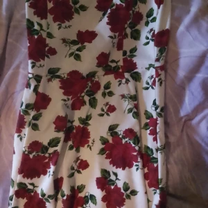 Vit klänning med röda blommor - Supersöt vit kortklänning med stora röda blommor och gröna blad. Klänningen har ärmlös design med breda axelband och en lätt utställd kjol som ger en skön och ungdomlig look. Perfekt för dig som gillar färgstarka mönster och vill sticka ut.