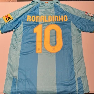 Barcelona Ronaldinho 10 Nike tröja - Säljer en ikonisk Barcelona fotbollströja med Ronaldinho och nummer 10 på ryggen. Tröjan är randig i ljusblå och blå toner, med gula detaljer och klubbmärke. Nike-logga och Unicef-tryck på bröstet. Tillverkad i lätt och ventilerande polyester. Replika, endast handtvätt gäller!