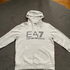 Vit hoodie från Emporio Armani EA7 - Snygg vit hoodie från Emporio Armani EA7 med stor logga framtill. Tröjan har huva med dragsko, känguruficka och långa ärmar. Tillverkad i mjuk bomull som känns skön mot huden. Perfekt för en clean och stilren look.