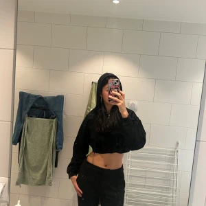 Svart croppad hoodie med dragkedja - Svart croppad hoodie med huva och snörning. Tröjan har en snygg silverfärgad half zip-dragkedja med ringdetalj framtill. Oversized passform och långa ärmar. Perfekt till jeans eller mjukisbyxor för en chill look.