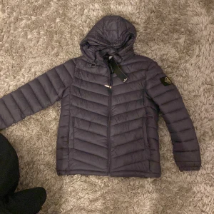 Svart dunjacka från Stone Island - Säljer en svart dunjacka från Stone Island med huva och klassisk patch på ärmen. Jackan har dragkedja framtill, två sidofickor och quiltad design. Perfekt för kalla dagar och riktigt snygg streetstil.