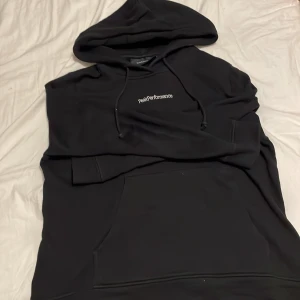 Svart hoodie från Peak Performance - Svart hoodie från Peak Performance med vit logga på bröstet. Klassisk modell med huva och dragsko samt stor magficka framtill. Mjuk insida och ribbade muddar vid ärmslut och nederkant. Perfekt för en avslappnad och sportig stil.
