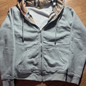 Grå hoodie från Burberry med dragkedja - Grå hoodie från Burberry med klassiskt rutigt foder i huvan och insidan. Tröjan har broderad logga på bröstet, snörning i huvan, två stora fickor framtill och dragkedja hela vägen. Perfekt för dig som gillar stilren streetwear med lyxig touch.