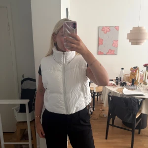 Vit dunväst från Bershka - Säljer en snygg vit dunväst från Bershka med hög krage och dragkedja framtill. Västen har en quiltad look och dragsko i nederkant för att justera passformen. Perfekt att slänga över en hoodie eller t-shirt för en chill vibe.