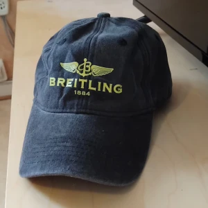 Svart keps från Breitling - Snygg svart keps från Breitling med gul broderad logga och text framtill. Klassisk böjd skärm och justerbar passform bak. Tillverkad i mjuk bomull som ger en skön känsla och cool look. Perfekt för dig som gillar stilrena accessoarer med tydlig branding.