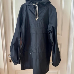 Svart anorak  från Vammas - Svart vindjacka från Vammas med huva och snörning i halsen. Jackan har lång ärm, knappar framtill och detaljer med metallöljetter på ärmarna. Tillverkad i slitstark bomull, perfekt för blåsiga dagar. Snygg och enkel design som funkar till många stilar.