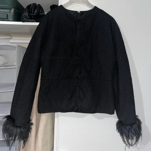 Svart jacka med snörning och fuskpäls - Svart jacka med cool snörning framtill och långa ärmar med fuskpälsdetaljer vid ärmsluten. Jackan har en rak passform och är tillverkad i ett strukturerat tyg som ger en lyxig känsla. Perfekt för dig som vill sticka ut med en unik look.