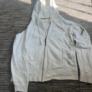 Vit hoodie från Acqua Limone med dragkedja - Vit hoodie från Acqua Limone med dragkedja framtill, huva och ribbade muddar. Stor logga med gul citron på ryggen och tryck på ena ärmen. Tillverkad i 100% bomull och har en ficka med dragkedja på ärmen. Perfekt för chill dagar. Finns lite fläckar här o där men skulle gissa på att det går bort i tvätten då den har luggit i men garderob ett tag. storlek M passar upp till 190cm. köpt för 1300kr på deras hemsida men har knappt använt. 
