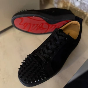 Christian Louboutin svarta sneakers med nitar - Säljer ett par svarta sneakers från Christian Louboutin med ikonisk röd sula och coola svarta nitar på tån. Skorna har snörning och är gjorda i mocka och skinn. Perfekta för dig som vill sticka ut med en lyxig och edgy stil.