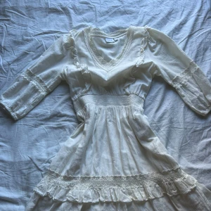 Vit spetsklänning från Vila - Romantisk vit klänning från Vila med v-ringning, spetsdetaljer och volanger. Klänningen har trekvartsärm och markerad midja med smock. Tillverkad i ett lätt material som känns luftigt och somrigt. Perfekt för dig som gillar en feminin och bohemisk stil. Använd 1 gång.