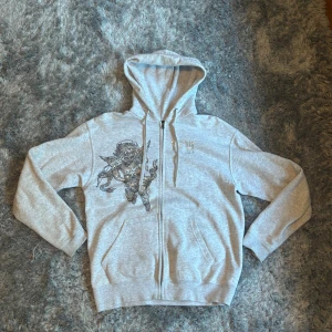 Grå bvaldi rhinestone zip up  - En riktigt fet Ljusgrå hoodie från Bvadi med dragkedja och huva. Framsidan har ett coolt tryck av en beväpnad ängel i svart. använt denna zip up en gång bara 