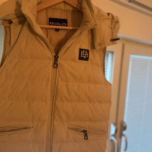 Beige manchester dunväst från Bvaldi  - Snygg beige dunväst från Bvaldi i manchester med diskreta ränder. Nypris 1800 och den är i fint skick utan defekter. Pris går att diskutera, skriv vid fler funderingar.