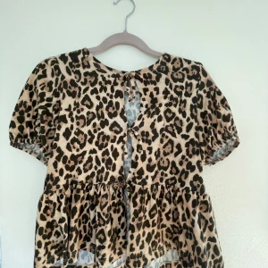 Leopardmönstrad blus från Gina Tricot - Säljer en superfin blus från Gina Tricot i leopardmönster. Toppen har korta puffärmar, volang nertill och två knytningar framtill som ger en snygg detalj. Färgerna går i beige och brunt med svarta inslag. Perfekt för dig som vill sticka ut lite extra.