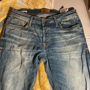 Blå jeans från Jack & Jones, stl 34/32 - Säljer ett par klassiska blå jeans från Jack & Jones i storlek 34/32. Jeansen har en snygg tvättad look med ljusa partier och kontrastsömmar. Femficksmodell med läderpatch bak i midjan. Perfekta till en avslappnad stil.