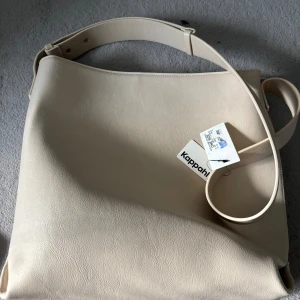 Beige axelväska från Kappahl - Stilren beige axelväska från Kappahl i syntetmaterial. Väskan har justerbart axelband, dragkedja upptill och flera fack inuti för smart organisering. Perfekt för dig som vill ha en clean och modern look till vardags.