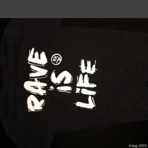 Svart Rave Culture T-shirt - Svart Rave Culture t-shirt från artisten W&W med vit tryckt text 'RAVE IS LIFE' på framsidan och coolt grafiskt mönster på baksidan. Skön passform och klassisk rund hals. Perfekt för dig som gillar streetwear och rave-vibes.